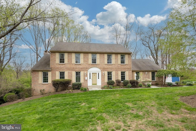 colonial-style house featuring a chimney, a front lawn, brick siding, and a shingled roof
