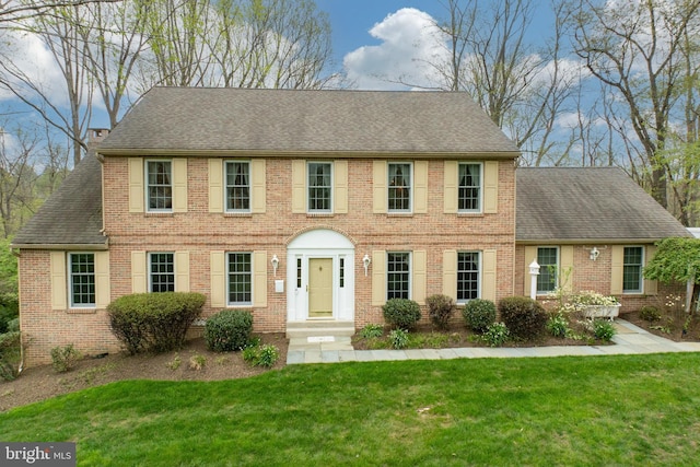 colonial house featuring a front lawn, brick siding, and roof with shingles