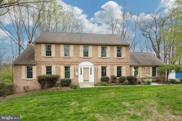 colonial house featuring a chimney, brick siding, a shingled roof, and a front yard