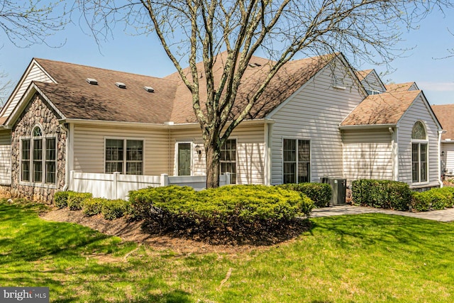 view of front of property with fence, stone siding, a front yard, roof with shingles, and central air condition unit