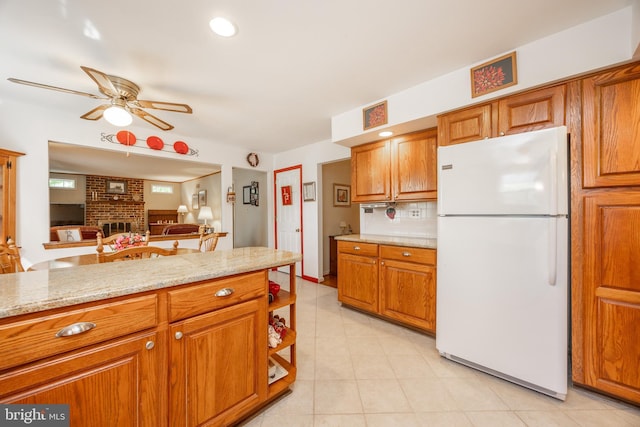 kitchen featuring ceiling fan, brown cabinetry, a brick fireplace, light tile patterned floors, and freestanding refrigerator