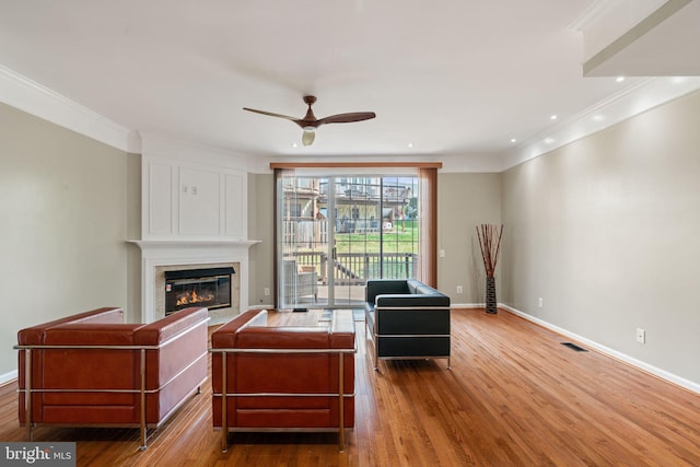 living area featuring crown molding, ceiling fan, a glass covered fireplace, baseboards, and wood finished floors
