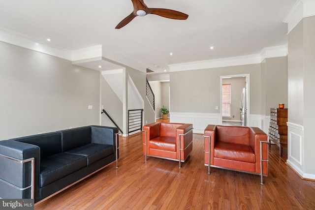 living area with crown molding, stairway, a wainscoted wall, hardwood / wood-style flooring, and a ceiling fan
