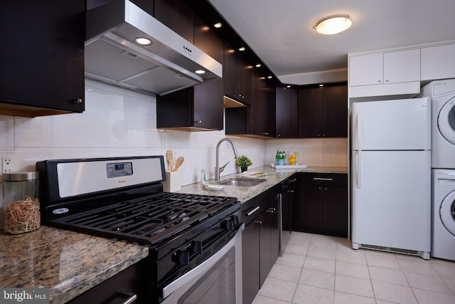 kitchen with a sink, stainless steel range with gas stovetop, stacked washing maching and dryer, freestanding refrigerator, and range hood