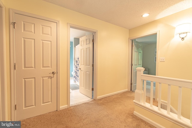 hall with a textured ceiling, light colored carpet, baseboards, and an upstairs landing