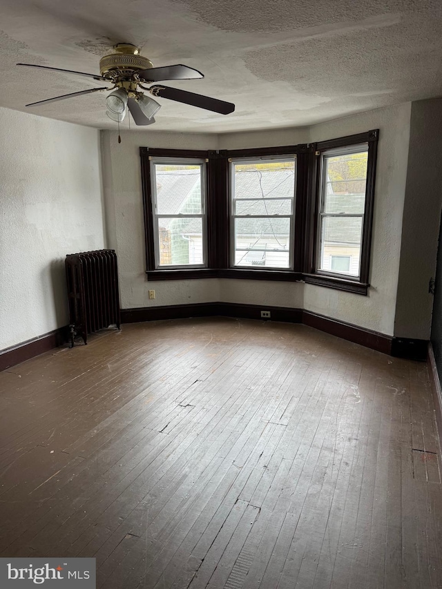 empty room featuring radiator heating unit, wood-type flooring, baseboards, ceiling fan, and a textured ceiling