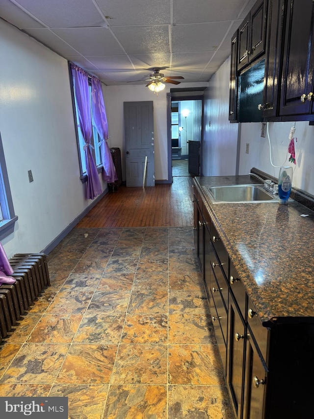 kitchen with a paneled ceiling, a ceiling fan, a sink, dark countertops, and baseboards