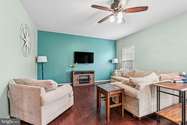 living area featuring baseboards, a ceiling fan, dark wood-type flooring, and a glass covered fireplace