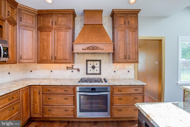 kitchen with light stone counters, custom exhaust hood, brown cabinets, and stainless steel appliances