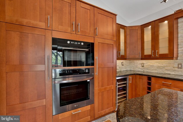 kitchen featuring dark stone countertops, brown cabinetry, beverage cooler, stainless steel double oven, and backsplash
