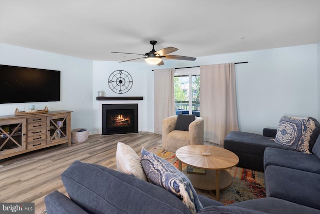 living room with a ceiling fan, a warm lit fireplace, and wood finished floors