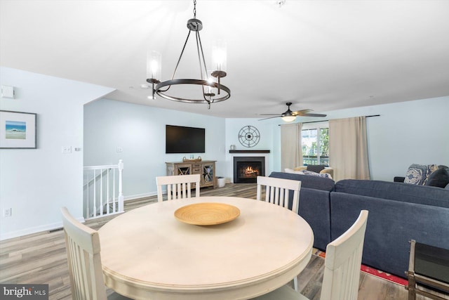 dining space with baseboards, light wood-style flooring, ceiling fan with notable chandelier, and a lit fireplace