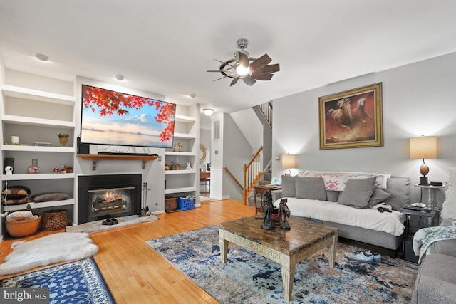 living room with ceiling fan, built in features, a glass covered fireplace, stairs, and wood finished floors