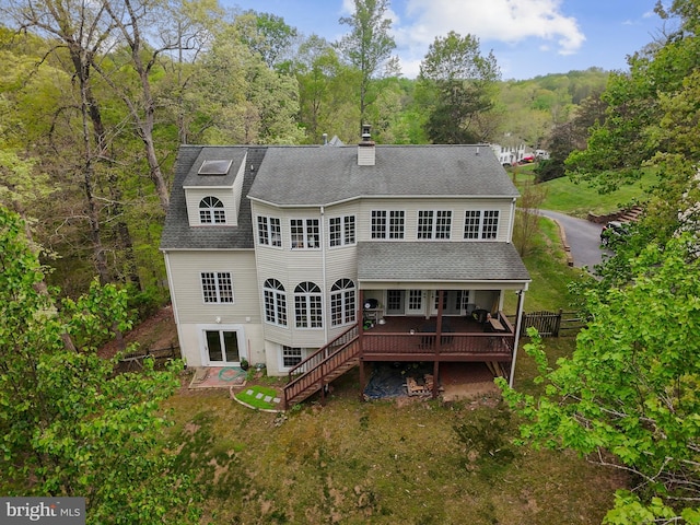 rear view of house with a deck, a chimney, a view of trees, and roof with shingles