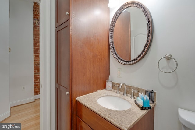 bathroom featuring wood finished floors, toilet, vanity, baseboards, and brick wall