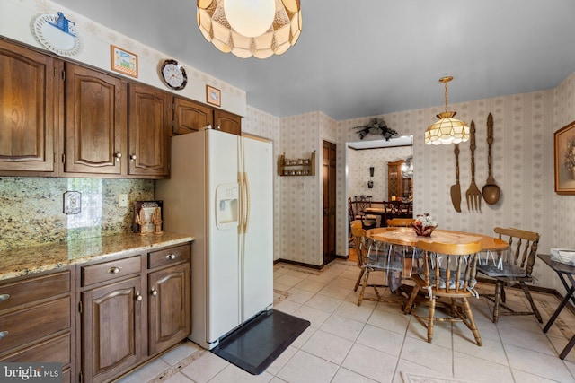 kitchen with white refrigerator with ice dispenser, wallpapered walls, light tile patterned floors, hanging light fixtures, and light stone countertops
