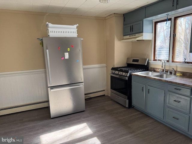 kitchen with a sink, a baseboard radiator, stainless steel appliances, a wainscoted wall, and under cabinet range hood