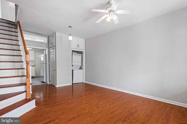 unfurnished living room featuring a ceiling fan, wood finished floors, stairs, stacked washer / drying machine, and baseboards