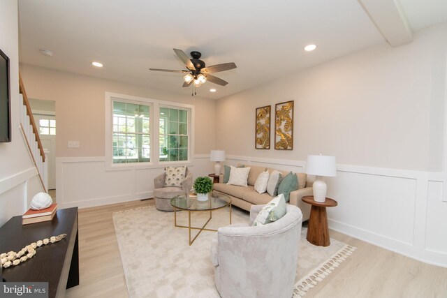 living area with a decorative wall, wainscoting, ceiling fan, light wood finished floors, and stairway