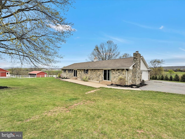 back of house featuring driveway, stone siding, a chimney, fence, and a lawn