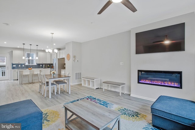 living room with visible vents, ceiling fan with notable chandelier, a glass covered fireplace, light wood-style floors, and recessed lighting