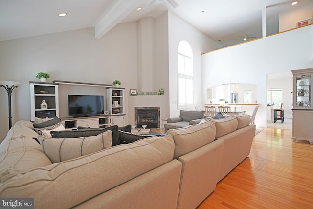 living area with ceiling fan, high vaulted ceiling, light wood-style flooring, beam ceiling, and a glass covered fireplace