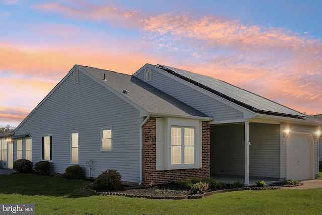 property exterior at dusk with a garage, brick siding, a yard, roof with shingles, and roof mounted solar panels
