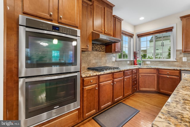 kitchen featuring appliances with stainless steel finishes, under cabinet range hood, a sink, light wood-type flooring, and tasteful backsplash