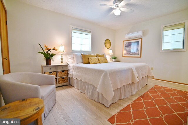 bedroom with baseboards, light wood-style flooring, ceiling fan, and a wall mounted AC
