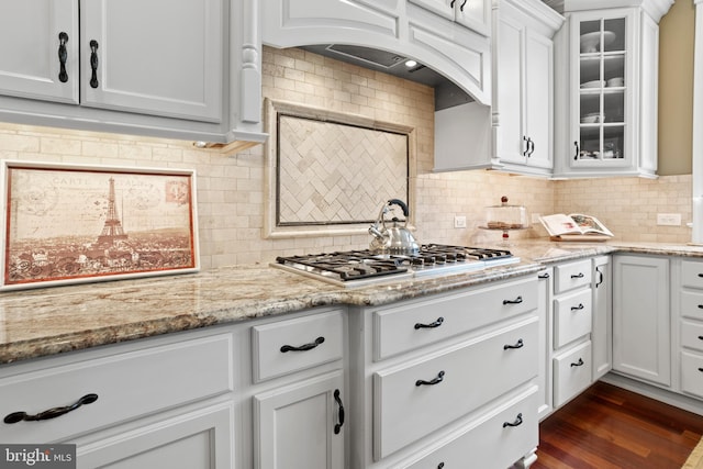 kitchen with stainless steel gas cooktop, white cabinetry, custom range hood, backsplash, and glass insert cabinets
