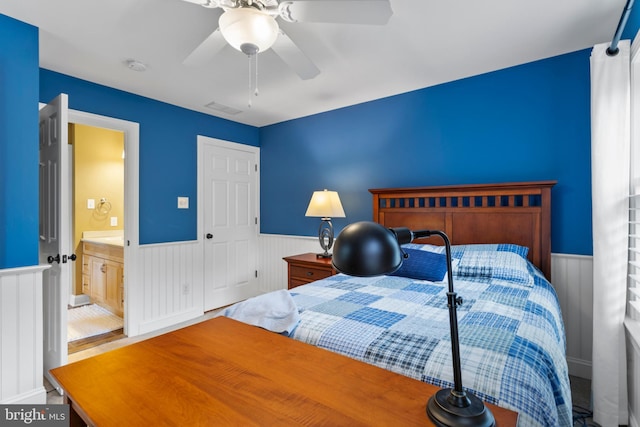 bedroom featuring ceiling fan, visible vents, ensuite bathroom, and a wainscoted wall