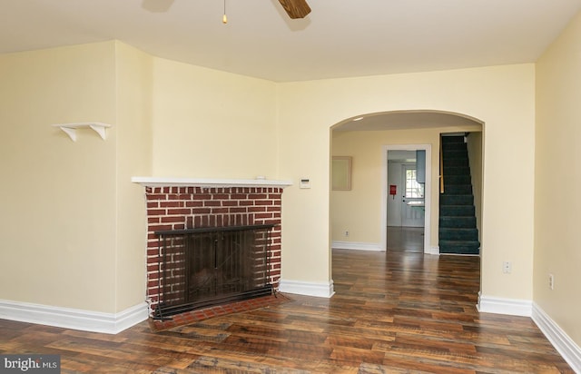 unfurnished living room with wood finished floors, arched walkways, baseboards, a brick fireplace, and ceiling fan