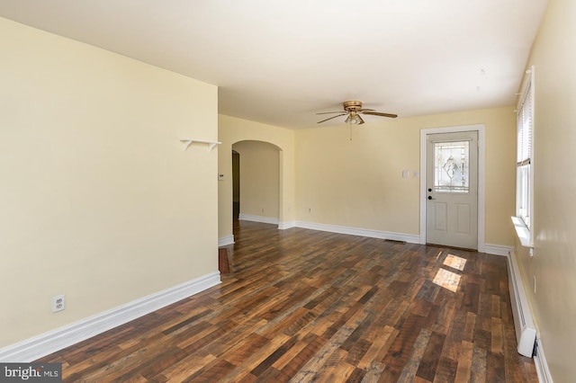 unfurnished living room with dark wood finished floors, arched walkways, a baseboard heating unit, baseboards, and ceiling fan