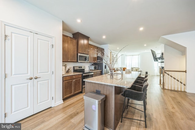 kitchen with a kitchen island with sink, light stone countertops, a kitchen bar, light wood-type flooring, and stainless steel appliances