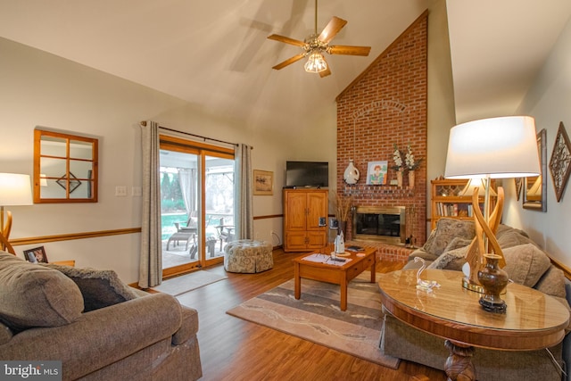 living room featuring high vaulted ceiling, a brick fireplace, ceiling fan, and wood finished floors