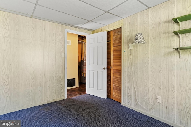 interior space featuring wooden walls, water heater, dark colored carpet, a drop ceiling, and visible vents