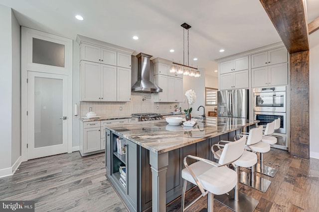 kitchen with a breakfast bar area, stainless steel appliances, light stone countertops, wall chimney exhaust hood, and wood finished floors