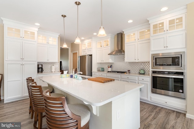 kitchen featuring wall chimney range hood, a kitchen island with sink, stainless steel appliances, light wood-type flooring, and tasteful backsplash
