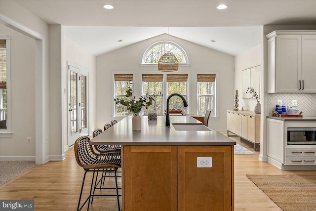 kitchen featuring a center island with sink, a sink, stainless steel microwave, lofted ceiling, and light wood-style flooring