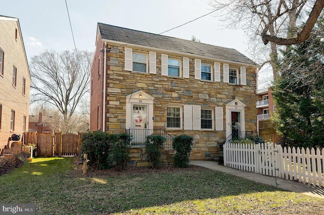 colonial inspired home with roof with shingles, fence, stone siding, and a front lawn