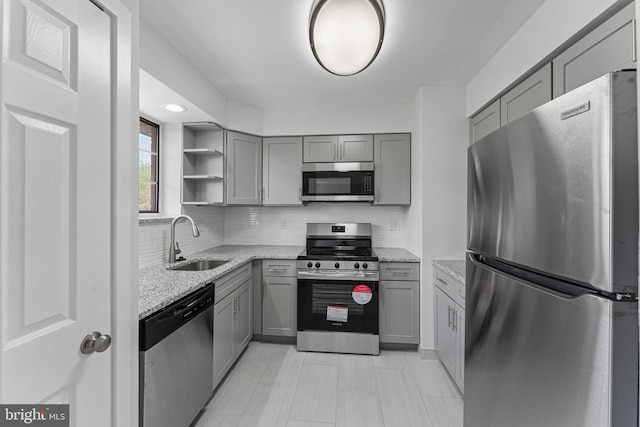 kitchen with open shelves, gray cabinets, light stone counters, a sink, and appliances with stainless steel finishes
