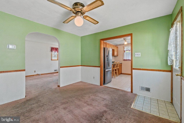 carpeted empty room with arched walkways, ceiling fan, wainscoting, and visible vents