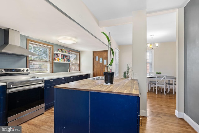 kitchen with wall chimney range hood, blue cabinetry, stainless steel range with electric stovetop, a sink, and wood counters
