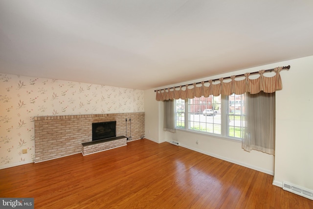 unfurnished living room featuring a brick fireplace, baseboards, wallpapered walls, wood finished floors, and visible vents