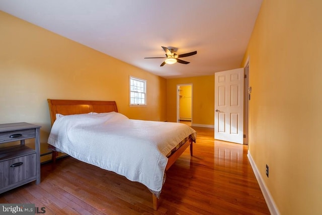 bedroom with a baseboard radiator, wood finished floors, a spacious closet, ceiling fan, and baseboards
