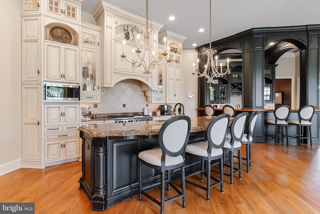 kitchen with dark stone countertops, stainless steel microwave, decorative backsplash, a kitchen island with sink, and a notable chandelier