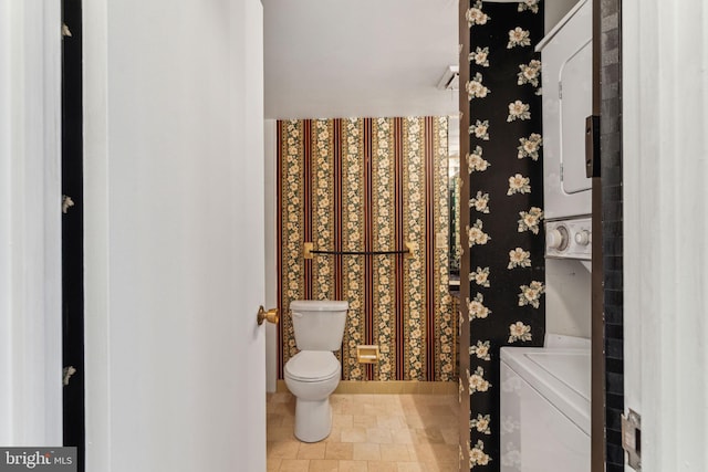 bathroom featuring stacked washer and clothes dryer, toilet, and wallpapered walls