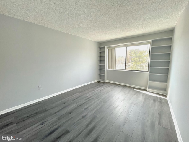 empty room with a textured ceiling, built in shelves, baseboards, and dark wood-style flooring