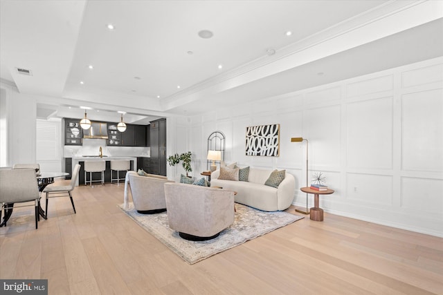 living area with a raised ceiling, visible vents, a decorative wall, crown molding, and light wood-style flooring