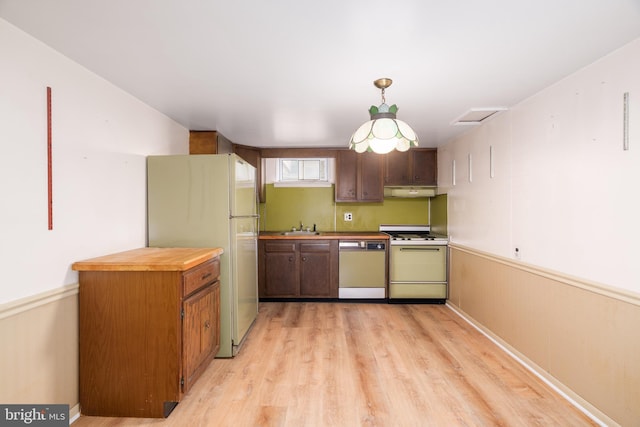 kitchen featuring a sink, under cabinet range hood, light wood-style floors, white appliances, and hanging light fixtures